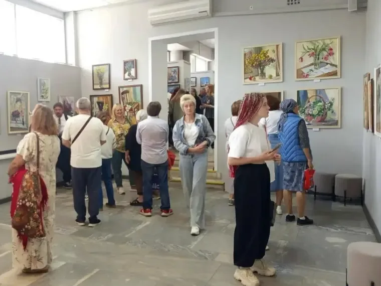 Посетители рассматривают картины на выставке в зале имени К. Малевича, окруженные живописными работами Веры Мазаевой.