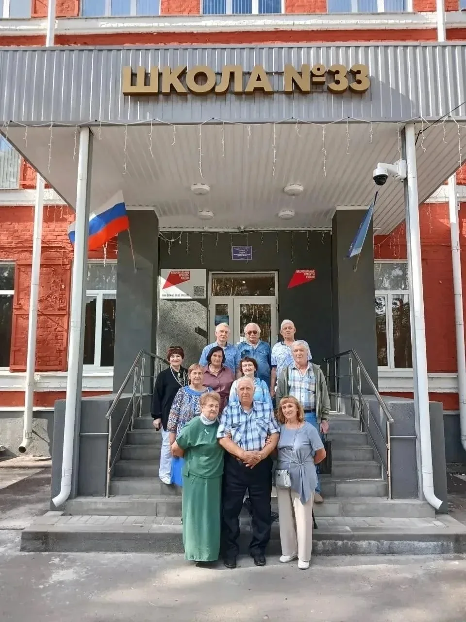 Группа пожилых людей на фоне здания школы, обсуждающих свои школьные годы и делящихся воспоминаниями.